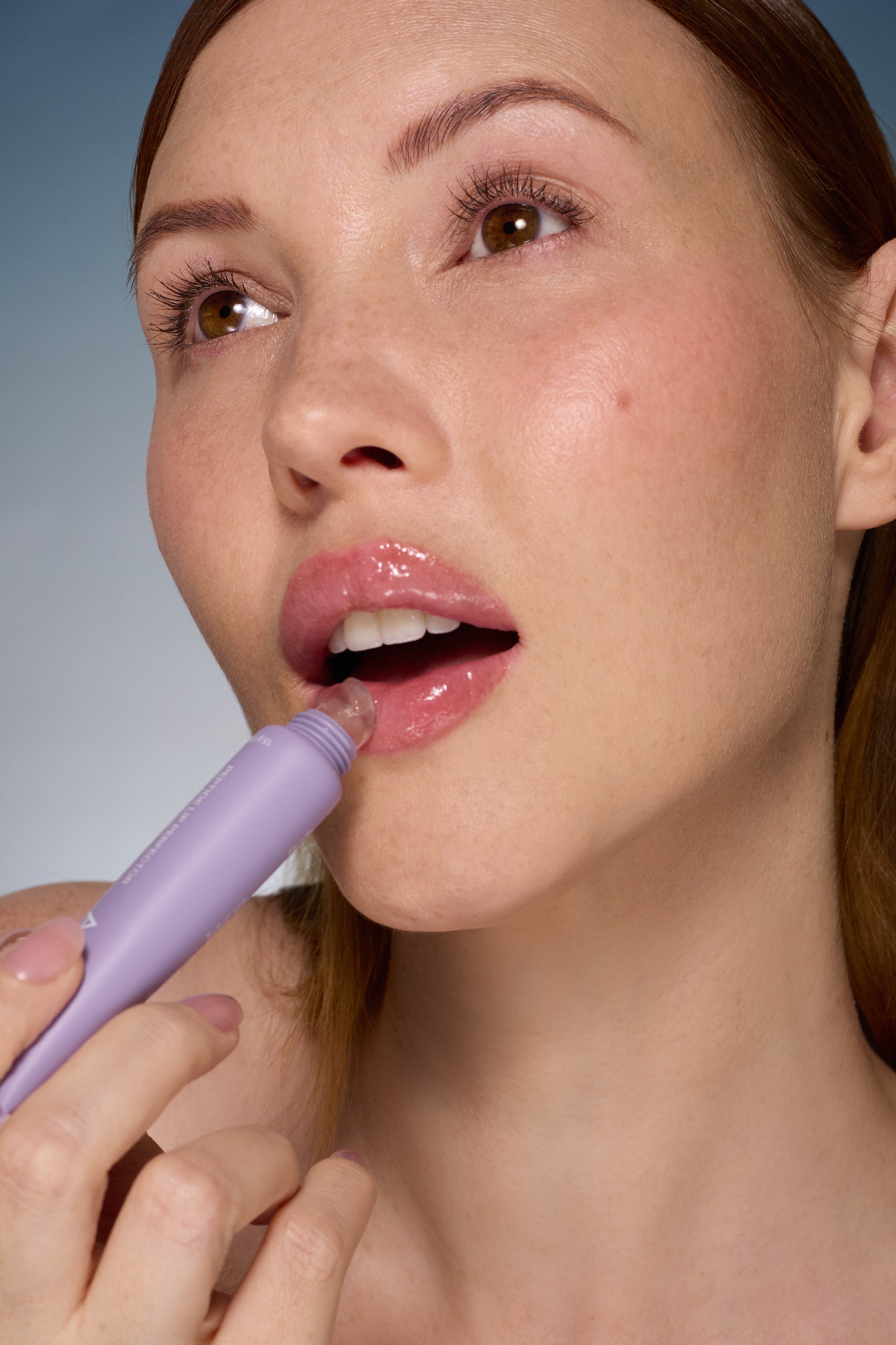 Women applying Alpha-H Peptide Lip Perfector lip balm to her lips. Purple applicator, light blue background.